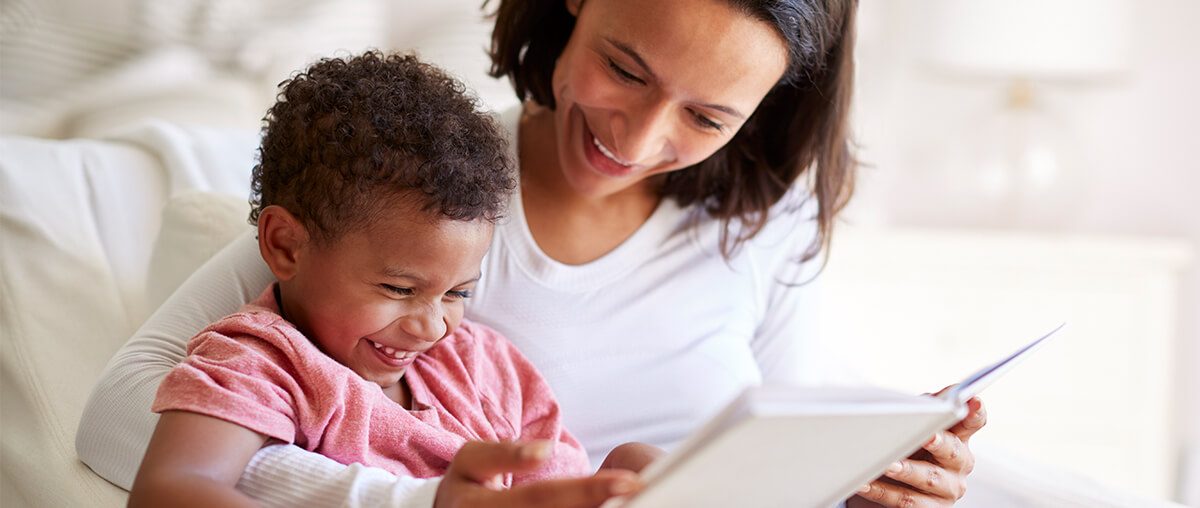 Close up of mixed race young adult mother sitting in an armchair reading a book with her three year old son on her knee, laughing.